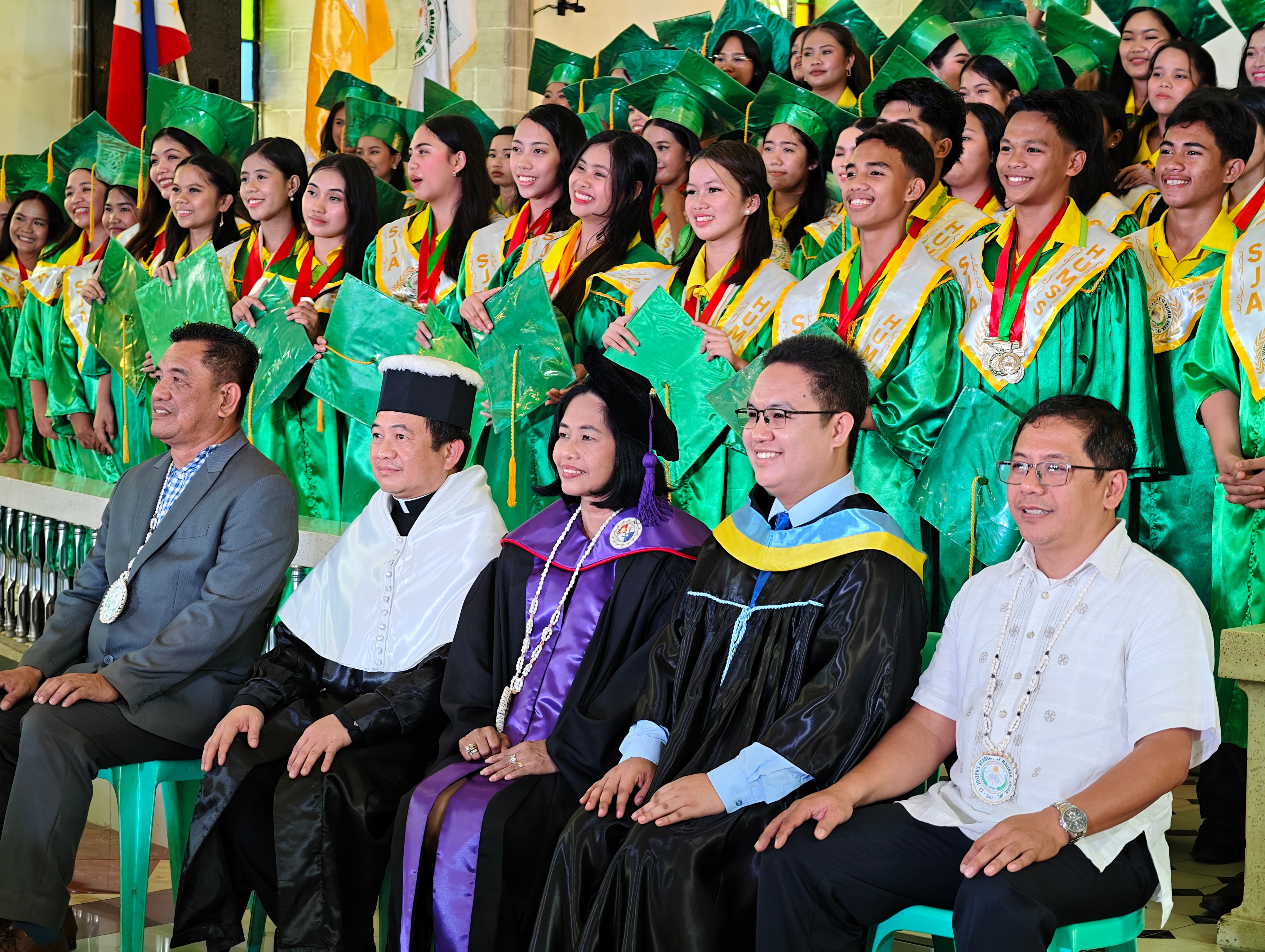 Graduating Guardians in green and gold gowns with faculty and the parish priest at commencement.