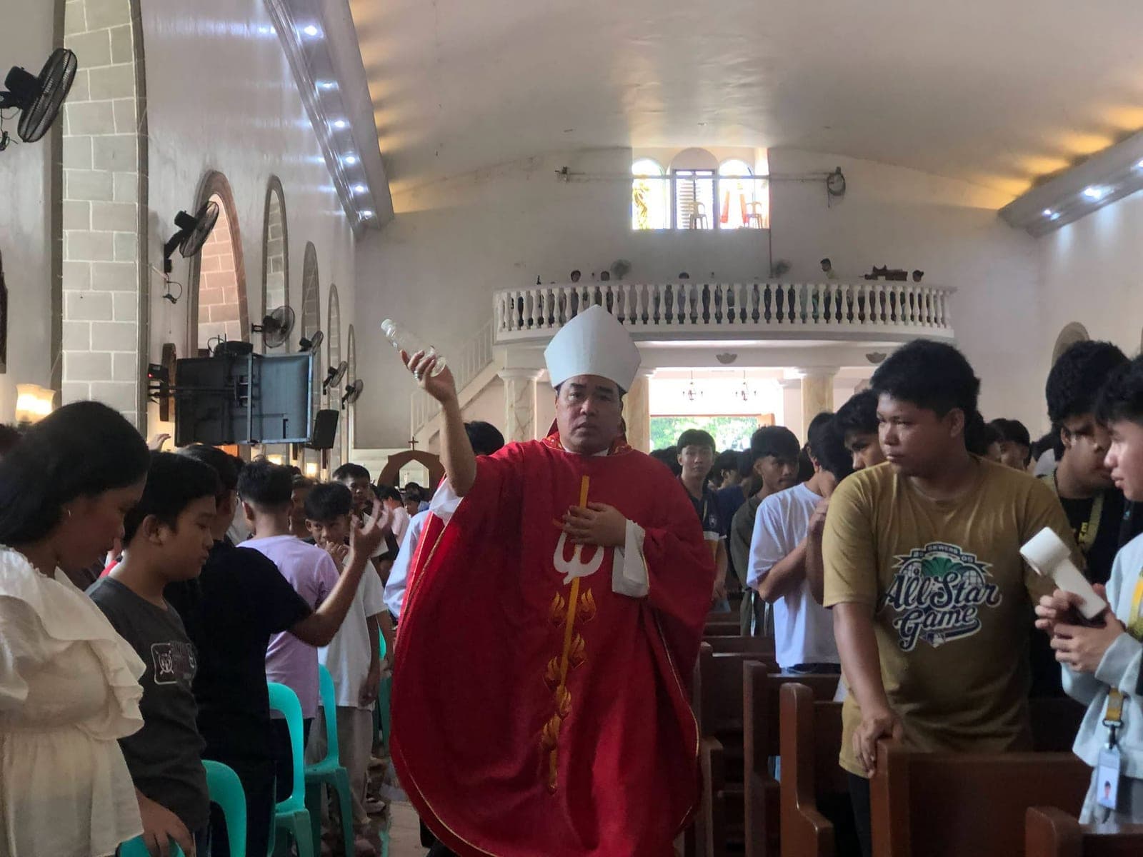 A bishop in red vestments walks down the church aisle sprinkling holy water while students stand at the pews on both sides