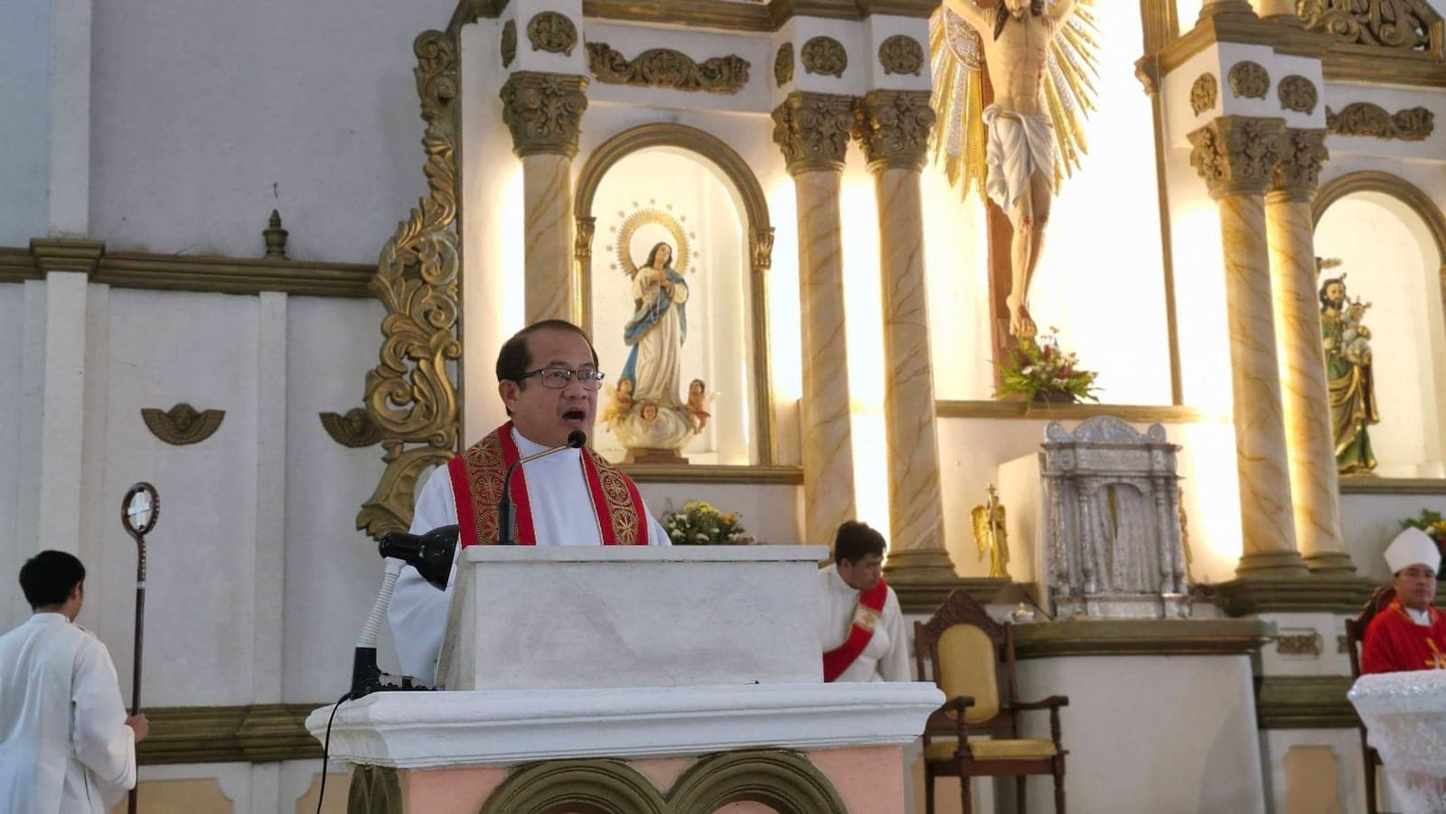 A priest in a red stole speaks from the pulpit, with the church's gilded altar and religious statues behind him